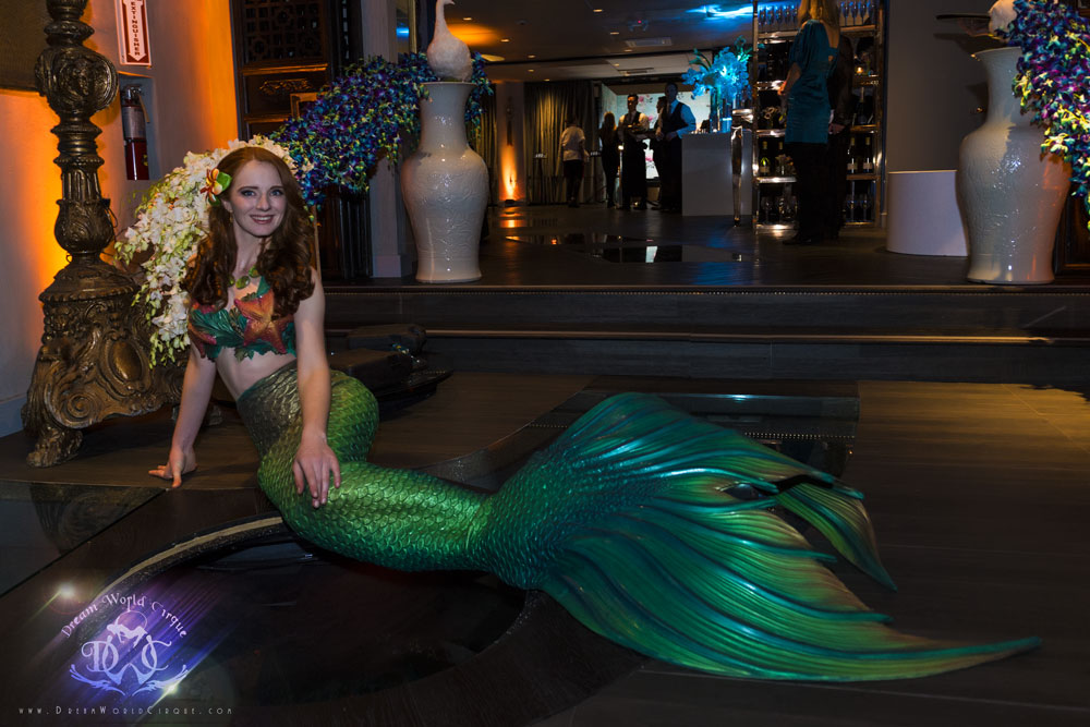 A beautiful red haired mermaid with vibrant green and turquoise tail lounges by an indoor pool