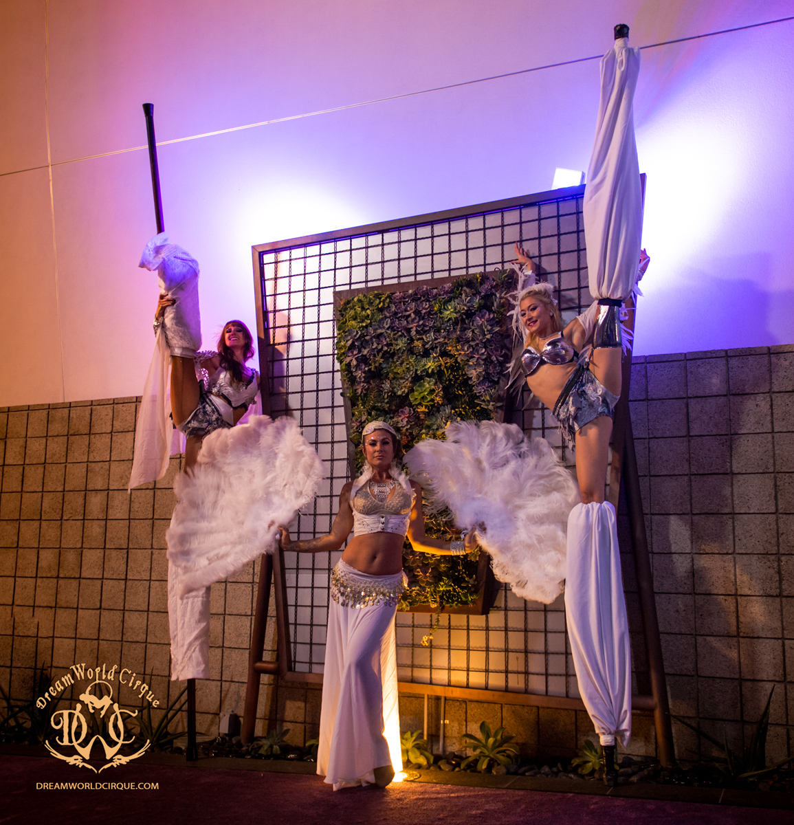 two tall long legged stilt dancers pose with legs lifted high in front of a floral arrangement with a elegant Feather Fan Dancer between them